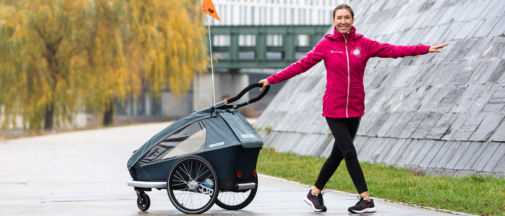 Woman performing cross step with stretched out arm while pushing Croozer bike trailer in autumn setting